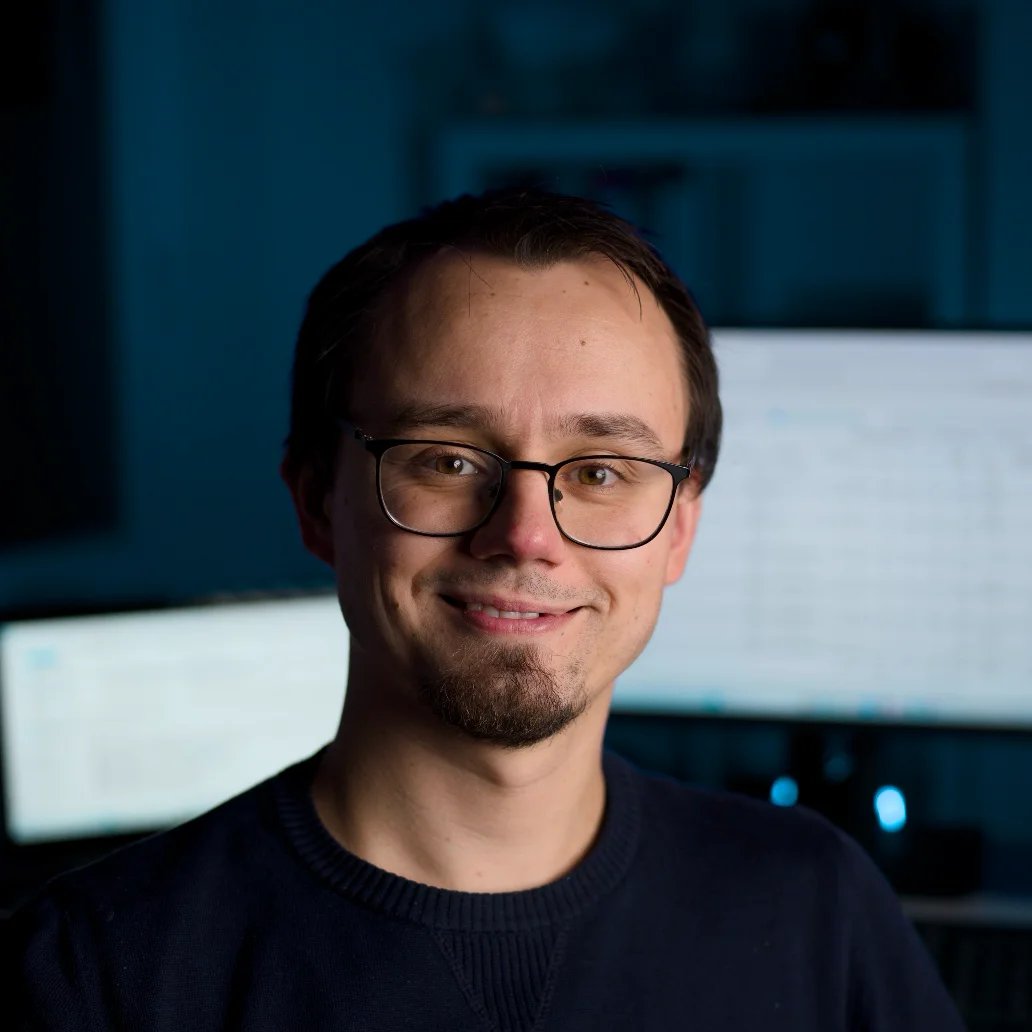 A person with glasses and a goatee smiles in a dimly lit room, with computer screens glowing in the background.
