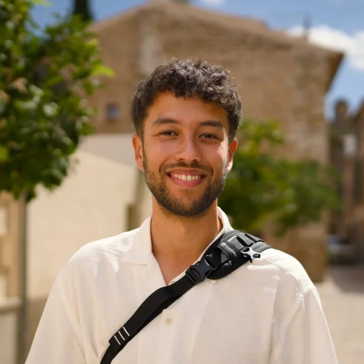 Smiling man with curly hair wearing a white shirt and black shoulder bag, standing outdoors with blurred buildings and trees in the background.