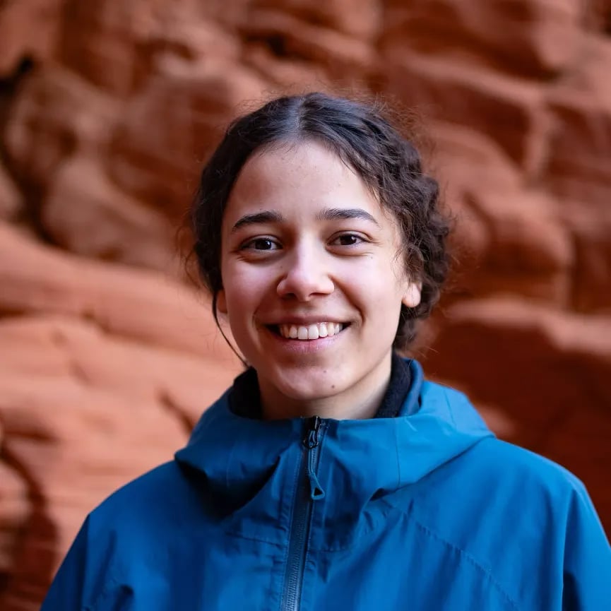 A person in a blue jacket smiles against a backdrop of red rock formations.
